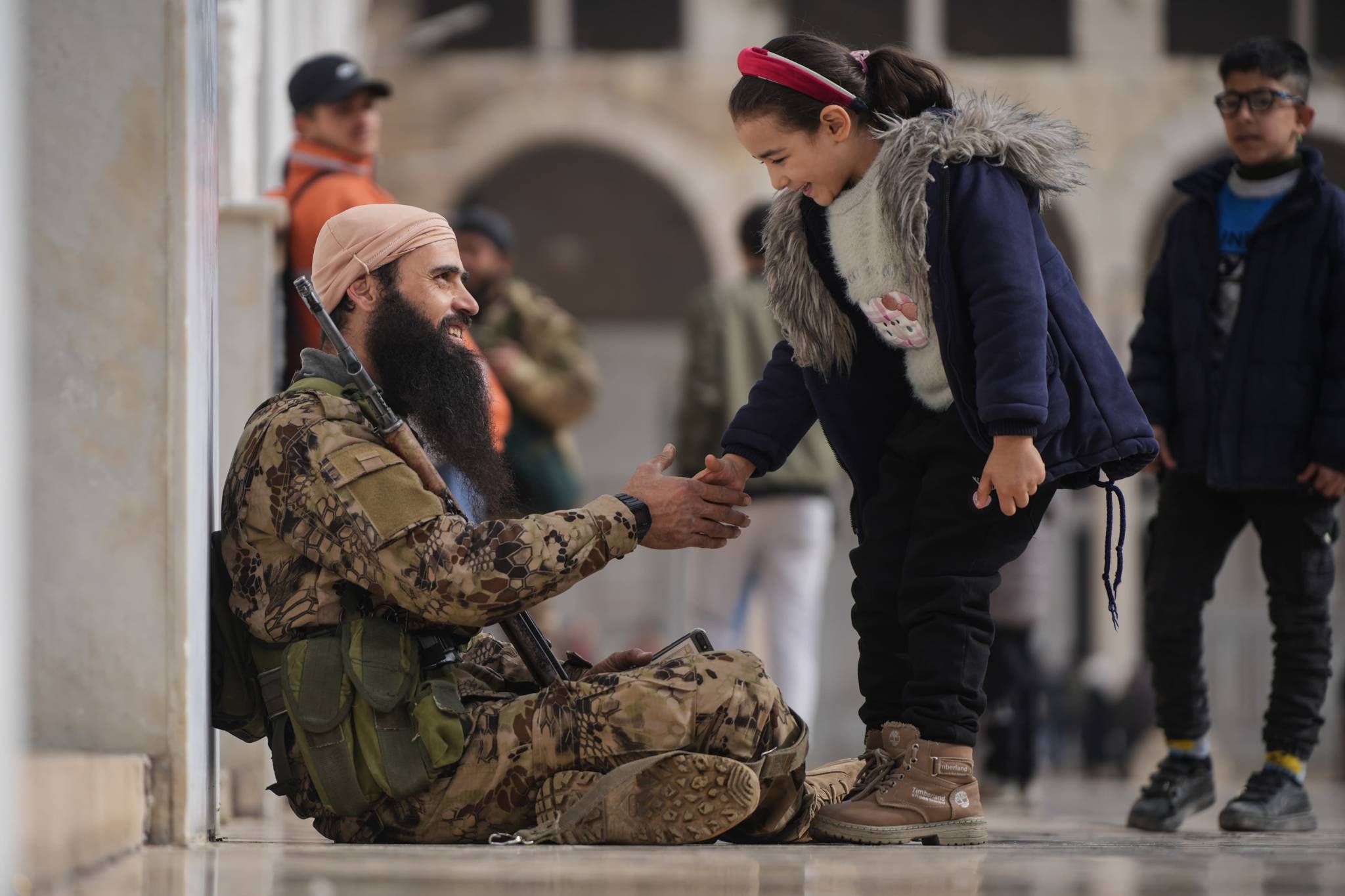 Both smiling, a little girl and a rebel fighter shake hands as he rests on a corner in a city that now has a very different attitude towards those who patrol the streets. (Credit: Mohammad Yassin/L'Orient Today)